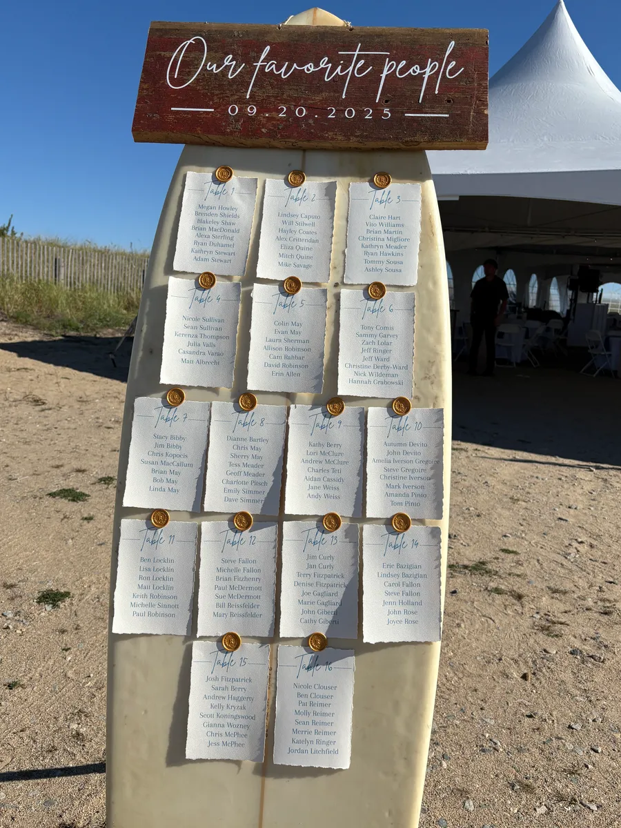 Beach wedding seating chart with sixteen table cards pinned to a surfboard under a wooden sign that reads our favorite people.