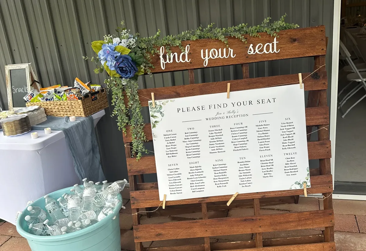 Wedding seating chart sign on a wooden pallet listing guest names by table number, decorated with eucalyptus and blue flowers at a reception entrance.